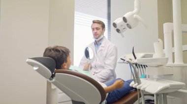 Neşeli küçük bir çocuk, arkadaş canlısı erkek dişçisi eşliğinde modern bir dişçi koltuğunda otururken kendine güvenle ışıldıyor. Gelişmiş araçlar ve parlak ışıklandırma ile dolu diş sağlığı kliniğinin sıcak atmosferi, paylaşılan güven ve ilgiyi vurgular
