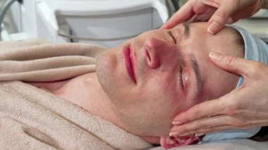Yakışıklı bir adam gözleri kapalı rahatlar, sakin bir kaplıca merkezinde rahatlatıcı bir yüz masajı yaptırır. Yetenekli masözlerin nazik dokunuşları cilt rutinini geliştirir. Huzurlu bir atmosferde rahatlama ve gençleşme sağlar.