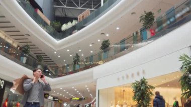 Şık bir genç adam hareketli bir alışveriş merkezinde canlı bir telefon görüşmesi yapıyor, omzunda zahmetsizce alışveriş torbalarını dengeliyor. Modern dekor ve yemyeşil ile dolu canlı atmosfer, onun alışveriş heyecanını artırır.