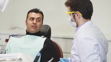 Olgun bir İspanyol diş hekimi, parlak, steril bir klinikte diş ağrısından bahsederken gözle görülür bir şekilde endişeli bir şekilde dişçi koltuğunda oturur. Koruyucu giysiler giyen dişçi, erkeğin ağzını dikkatle inceler.
