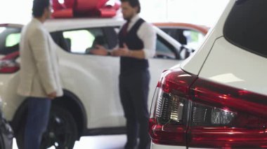 Gösterişli beyaz bir araba ön planda belirgin bir şekilde dururken, profesyonel bir otomobil satıcısı olgun bir adama arabanın kapısını açarak yardım ediyor. Bu ilgi çekici sahne, yeni bir araç seçmenin heyecanını yakalıyor.