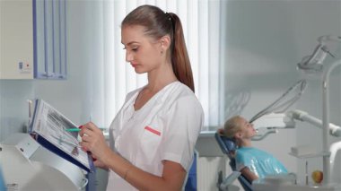 Gösterişli bir dişçi dolabında, odaklanmış beyaz bir dişçi elinde mavi bir kalem tutarken hasta kayıtlarını inceleyerek, kaliteli tedaviye olan bağlılığını gösteriyor. Arkasında, dişçi koltuğunda rahatça oturan ve tedaviye hazır rahat bir kadın hasta var.