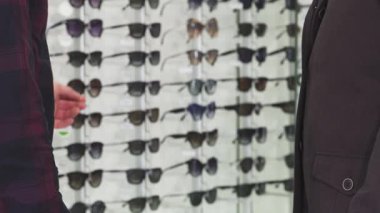 Yakın plan çekimde profesyonel bir optometrist ve erkek müşterisi sıkı bir tokalaşma içinde başarılı bir göz alımını kutluyor. Şık güneş gözlüklerinin arka planı modern bir yetenek katarak memnuniyet atmosferini arttırıyor.
