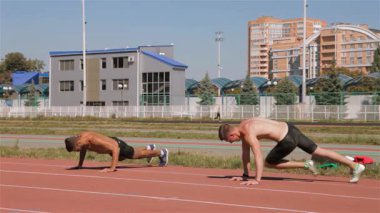 İki kararlı beyaz erkek sporcu stadyumda dinamik bir ısınma turuna katılıp atletizmlerini sergiliyorlar. Formda bir adam yüzükoyun yerde, yoğun bir antrenmana hazırlanırken bacaklarını büküyor, diğeri ise odaklandığı şeyi yansıtıyor.