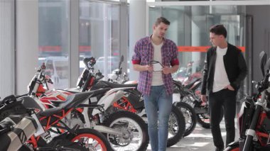 Kendine güvenen bir satıcı ve hevesli bir müşteri el sıkışırken dinamik bir sahne ortaya çıkıyor ve çarpıcı çapraz motosikletlerin arasında bir anlaşma imzalıyor. Işıldayan makineler ve canlı renklerle dolu parlak sergi salonu heyecan verici bir atmosfer yaratıyor.