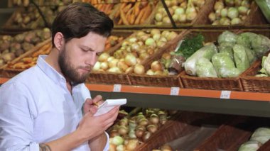Tarz sahibi beyaz bir adam, süpermarketteki canlı taze ürünler arasında defterine yazdığı gibi derin düşüncelere dalmış durumda. Elinde bir kalem ve hafif bir gülümsemeyle, renkli sebzelerle çevrili alışveriş listesini dikkatle hazırlar.
