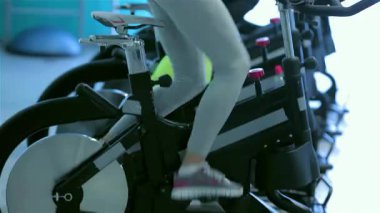 Ton bacaklı atletik bir kız, dört formda arkadaşı tarafından çevrelenmiş durgun bir bisikletin üzerinde güçlü bir şekilde pedal çeviriyor, hepsi yoğun bir çalışma içindeler. Enerjik spor salonu atmosferi sınırlarını zorlarken enerji ile uğulduyor.