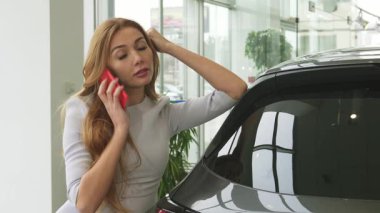 Modern bir araba galerisindeki lüks bir araca yaslanmış akıllı telefonuyla sohbet ederken neşeyle saçlarını dalgalandıran çarpıcı genç bir kadın. Parlak, havadar sergi salonu onun heyecanını ve lüks arabaların cazibesini yansıtıyor.