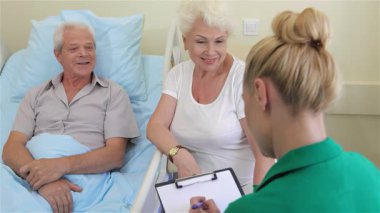 Şefkatli bir hemşire, kıdemli bir erkek hasta ve onun destekleyici karısıyla parlak bir hastane koğuşunda karşılaşır. Yeşil üniforma giymiş sarışın hemşire, elinde bir pano tutarken gri saçlı adam gülümsüyor ve sıcak bir bakım ortamı yaratıyor.