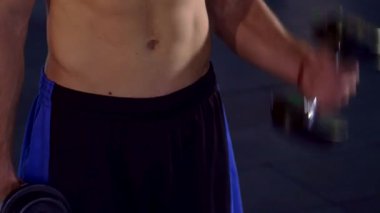 Odaklanmış genç bir beyaz atlet, etkileyici fiziğini sergileyerek spor salonunda pazılarını tutkuyla çalıştırıyor. Kamera, güçlü gövdesinin yakın çekimini çekiyor. Halterlerle farklı kıvrımlar sergiliyor ve vücudunun tanımını vurguluyor.