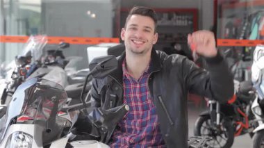 Neşeli esmer bir adam elinde motosiklet anahtarlarını sallıyor, galeride heyecan saçıyor. Şık bir deri ceket giyer, parlak beyaz bir motosikletin üzerine güvenle oturur, yeni maceraların heyecanını somutlaştırır.