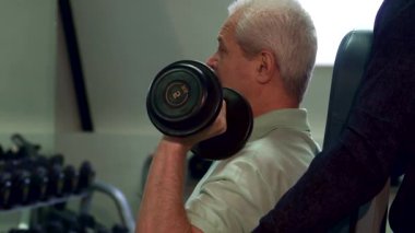 Kendini adamış genç bir Kafkasyalı antrenör, kıdemli bir müşteriye yardımcı oluyor. Güç antrenmanı sırasında düzgün bir form sağlıyor. Antrenörlerin yakın plan çekimleri, yaşlı adamların dirseklerini desteklerken, bakım ve bakım bağını vurgularken odaklanmış ifadelerini yakalar.