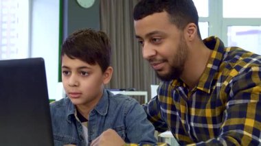 Özenli Afrikalı Amerikalı babası ona gülümseyerek rehberlik ederken, dikkatle dizüstü bilgisayarda esmer saçlı tiplere odaklanmış genç bir çocuk. Bu iç açıcı sahne bir bağ ve öğrenme anını yakalar, samimi ve modern bir şekilde bağlarını gösterir.