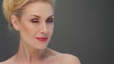 Çekici, sarışın bir kadın büyüleyici bir bakışla hayranlık duyuyor. Kusursuz cildi, yumuşak, gri bir zemin üzerinde parıldıyor. Bu portre, güzelliğin ve zarafetin özünü yakalıyor. Nazik gülümsemesini ve narin yüz hatlarını gözler önüne seriyor. Seyircileri davet ediyor.