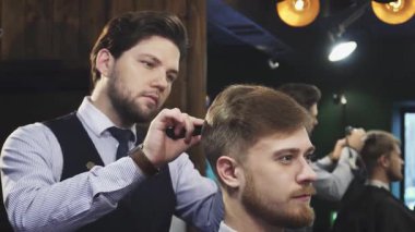 Ilık ışıkla süslenmiş şık bir berber dükkanında, yetenekli bir berber titizlikle genç ve yakışıklı bir adamın saçını tarar. Atmosfer güven ve rahatlama yayar. Bakım sanatını ve yeni saç kesiminin memnuniyetini gösterir.