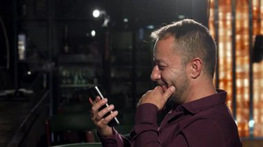 İki neşeli adam, biri şık sakallı ve gözlüklü, canlı bir bar ortamında kameraya neşeyle gülerken içkileriyle kadeh kaldırıyor. Yakışıklı, olgun bir adam arkadaşına heyecanlı bir şekilde akıllı telefonuyla bir şey gösteriyor.