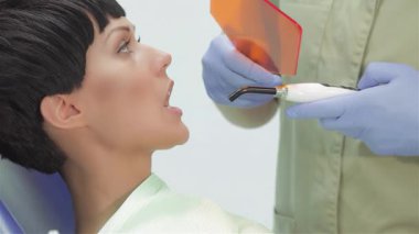 Parlak, modern bir dişçi kliniğinde, yetenekli bir dişçi, genç ve güzel bir kadının dişine ultraviyole fotopolimer mühür takar ve bu da optimal bakımı sağlar. Odaklanmış atmosfer diş tedavisinin hassasiyetini vurguluyor.