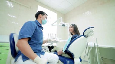 Modern bir diş kliniğinde, maskeli genç bir dişçi gülümseyen bir hastayla hevesle iletişim kurar. Birlikte, kameraya doğru dönerek, her ikisi de neşe dolu bir onay veriyorlar, pozitif enerji yayıyorlar ve diş bakımına güveniyorlar.