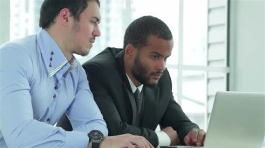 Biri şık takım elbiseli, diğeri şık gömlekli iki başarılı iş adamı, çağdaş bir ofis masasında dizüstü bilgisayarla birlikte çalışıyorlar. İfadeleri, profesyonelliğin özünü somutlaştıran kararlılık ve takım çalışmasını yansıtıyor.