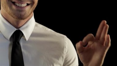 Gıcır gıcır beyaz gömlekli ve şık siyah kravatlı şık beyaz bir adam, eliyle iyi bir jest yaparken kendine güvenen bir gülümseme sergiliyor. Bu çarpıcı görüntü, kurumsal temalar ve iletişim için mükemmel bir pozitiflik ve profesyonellik yayıyor.