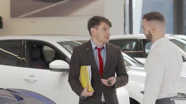 Şık bir takım elbise giymiş neşeli bir araba satıcısı, arabanın anahtarlarını aldıktan sonra heyecandan parlayan genç bir müşteriyle tokalaşıyor. Bu canlı araba galerisi otomotiv dünyasında güven ve ortaklığın özünü yakalıyor.