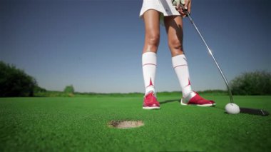 Şık kırmızı golf ayakkabıları giyen kararlı genç bir kadın, hassas bir vuruşla ustalıkla saklanıyor. Dikkatini topa nazikçe vurduğu bariz. Parlak güneş ışığı altında parlayan yeşil golf sahası enerjik bir enerji yaratıyor.