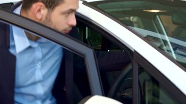 Bakımlı sakallı şık bir genç adam, modern bir arabada, düşünceli bir ifadeyi sergileyerek rahatça oturuyor. Parlak iç mekan ve parlak yüzeyler, yeni bir yolculuğa çıkmaya hazırlanırken heyecanını yansıtıyor.