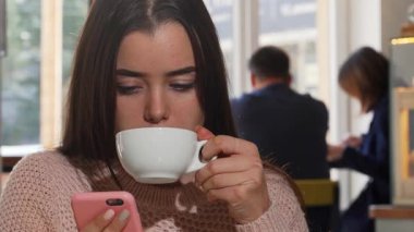 Çekici genç bir kadın sabah kahvesinin tadını çıkarıyor, mesajlar gönderirken dikkatle akıllı telefonuna odaklanıyor. Kafenin ılık atmosferi, arka planda hafif ışıklandırma ve bulanık figürlerle, rahatlama için mükemmel bir ortam yaratıyor.