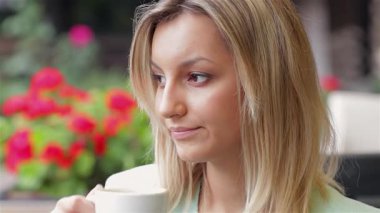 Şık bir iş kadını, çiçeklerle ve canlı bir şehir atmosferiyle çevrili açık bir kafede sıcak bir fincan kahvenin tadını çıkarıyor. Onun sakin tavrı ve düşünceli bakışı şehir hayatının koşuşturmacası ve telaşının arasında bir anlık sükuneti yansıtıyor.