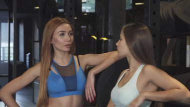 İki çarpıcı fitness meraklısı, biri dalgalı kızıl saçlı ve diğeri parlak siyah bukleli, modern bir spor salonunda güven içinde, neşe ve dostluk saçan pozlar veriyorlar. Canlı spor kıyafetleri ve olumlu jestleri dostluk ruhunu temsil ediyor.