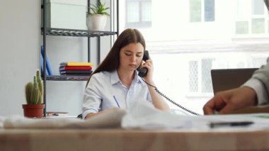 Genç, çekici bir bayan tasarımcı modern ofisinde bir telefon görüşmesine cevap verirken, bir meslektaşı ön plandaki bir giysi parçasını ölçüyor. Sahne, yaratıcılığın ve moda endüstrisindeki işbirliğinin dinamik enerjisini yakalıyor.