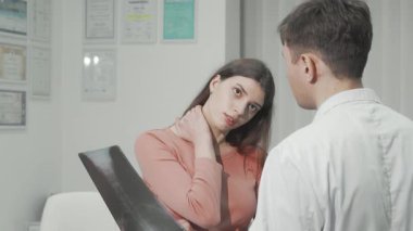 Endişeli bir kadın röntgen sonuçlarını parlak bir klinikte bir doktorla tartışırken boynunu sallıyor. Bu samimi an, sağlık hizmetlerinin önemini ve sürekli acıdan kurtulmanın yollarını vurguluyor.