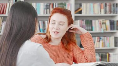 Biri uzun siyah saçlı, diğeri çarpıcı kızıl saçlı iki genç kadın, canlı bir üniversite kütüphanesinde renkli kitaplıklar arasında projelerini tartışıyorlar. İfadeleri coşku ve yoldaşlık yayıyor.