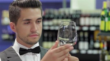 Şık yelekli ve papyonlu dengeli genç bir adam şarap yapımında uzmanlığını sergileyerek bir bardak kırmızı şarabı inceliyor. Zarif bir şarap rafıyla çevrelenmiş, odaklanmış ifadesi şarap tatma sanatında özgüven ve entelektüelliği yansıtıyor.