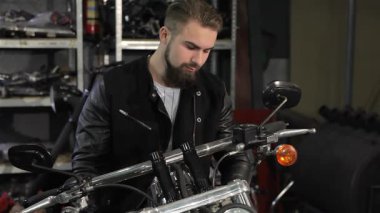 Genç, sakallı, parlak siyah deri ceketli beyaz bir adam helikopterinin gidonuna yaslanmış büyüleyici bir ifadeyle kameraya bakıyordu. Motorsiklet parçaları ile dolu canlı bir atölye arka planı,