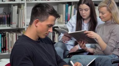 Çekici bir erkek öğrenci, ödevlerine dalmış, canlı bir kütüphane atmosferinde kameraya ışıldıyor. Öğrenci arkadaşlarla çevrelenmiş bu sahne, öğrenme sevinci ve pozitifliği yayarak akademik hayatın canlı enerjisini gözler önüne seriyor.