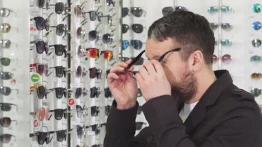 Optometrist dükkanında aynanın önünde son moda gözlüklerle deneyler yapan sakallı olgun bir adamın profil görüntüsü. Göz giysisinin bu canlı görüntüsü onun düşünceli ifadesini yansıtıyor. Mükemmel çifti bulmanın verdiği heyecanı gösteriyor.