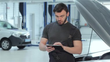 Yetenekli bir sakallı tamirci, parlak, modern bir garajda açık bir araba kaputunun yanında dururken dijital tableti dikkatle inceliyor. Parlak siyah bir üniforma giyen bu genç beyaz adam profesyonelliği ve uzmanlığı kusursuz bir şekilde harmanlıyor.