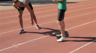 İki kararlı Kafkas erkek sporcu stadyumdaki canlı kırmızı pistte ısınıyor. Düşük açılı çekim, kalça esneme hareketleri sırasında odaklanmış hareketlerini yakalar, bir atlet öne eğilir ve diğeri de...