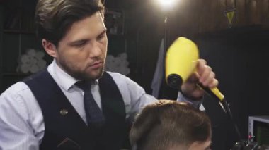 Şık, genç bir berber, şık bir yelek giymiş, müşterilerinin saçlarını ustalıkla kurutuyor, rahat bir berber dükkanında, profesyonellik ve yetenek yayıyor. Yumuşak ışıklandırma ve klasik dekor ile vurgulanan ılık ortam, samimi deneyimi güçlendirir.