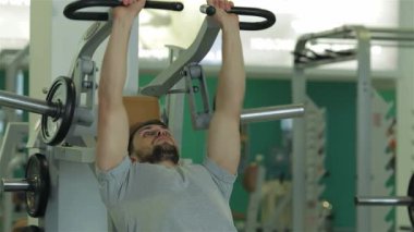 Güçlü vücutlu genç bir adam göğüs kaslarını yoğun bir şekilde çalıştırıyor, egzersiz aletlerini azimle kavrıyor. Modern fitness malzemeleriyle dolu canlı spor salonu atmosferi güçlendiriyor.