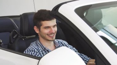 Neşeli genç bir adam, üstü açık arabasında otururken heyecandan kıpır kıpır oluyor. Arabanın anahtarlarını gururla gösteriyor. Bu canlı sahne yeni sahipliğin ve açık yolun özgürlüğünün heyecanını yakalıyor.