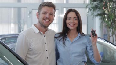 Galerideki yeni araba alımlarını kutlarken heyecan dolu bir çift. Genç beyaz adam, büyüleyici bir gülümseme ile, güzel esmer kız arkadaşının tuttuğu arabanın anahtarına hayran, parlak mutluluğu yayıyor.