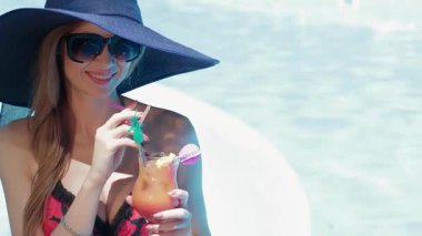 Parlak kırmızı mayo giymiş göz kamaştırıcı ince bir kadın, neşeli gülümsemesini çerçeveleyen koyu mavi şapkası ve tropikal bir kokteyl ile neşeli poz veriyor. Havuzun köpüklü suyu güneş ışığında parlıyor ve bu ferahlatıcı ortam için mükemmel bir zemin oluşturuyor.