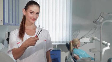 Kendine güvenen genç bir dişçi kadın cerrah dişçi aletlerini canlı kırmızı bir elma ile birlikte göstererek sağlık ve hijyeni sembolize ediyor. Kahverengi saçları arkadan bağlanmış ve beyaz bir ceket giyiyor. Modern bir dişçi kliniğinde duruyor.