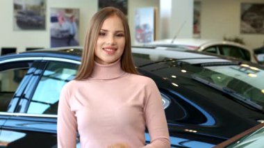 Kendine güvenen, kahverengi saçlı genç bir kadın lüks bir galeride araba anahtarını tutuyor ve yeni alışverişi için heyecanını sergiliyor. Arka planda, başarı ve memnuniyet atmosferini artıran çarpıcı siyah bir sedan yer alıyor.