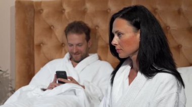 Rahat bir yatak odasında, olgun bir kadın sorunlu bir ifadeyle oturur, akıllı telefonuyla dikkati dağınık bir şekilde mesajlaşan kocasından uzağa bakar. Bu dokunaklı sahne ilişkilerindeki duygusal mesafeyi ve gerilimi yakalar.