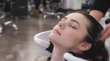 Huzurlu genç bir kadın şık bir spa salonunda gençleştirici şampuan banyosunun tadını çıkarıyor. Gözleri mutluluktan kapanıyor. Bir güzellik uzmanının becerikli elleri nazikçe kafa derisine masaj yapıyor, rahatlatıcı bir atmosfer yaratıyor kaliteli saç ürünleri ile dolu