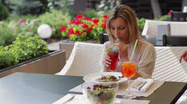 İki şık kadın canlı kokteyllerini yudumlarken çiçeklerle süslenmiş şık bir kafede gülüp sohbet ediyorlar. Hayat dolu atmosfer neşe ve dostluk saçıyor. Harika bir yaz tatili oluyor. Hoş sohbetlerle dolu.