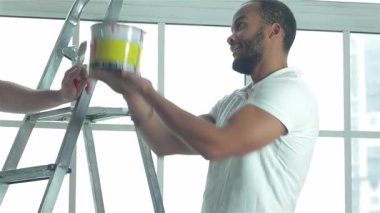 Parlak, havadar bir dairede, iki usta işçi son ölçümler ve tamiratlar için işbirliği yapıyor, yakınlarda bir merdiven ve canlı bir boya kovası var. Mekanı değiştirmeye hazırlanırken takım çalışmaları parlıyor, yenilenme ruhunu somutlaştırıyor ve